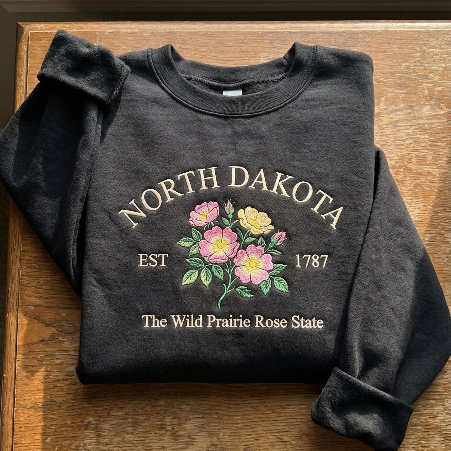 Charcoal black sweatshirt with detailed pink and yellow Wild Prairie Rose flower embroidery reading North Dakota EST 1787 The Wild Prairie Rose State