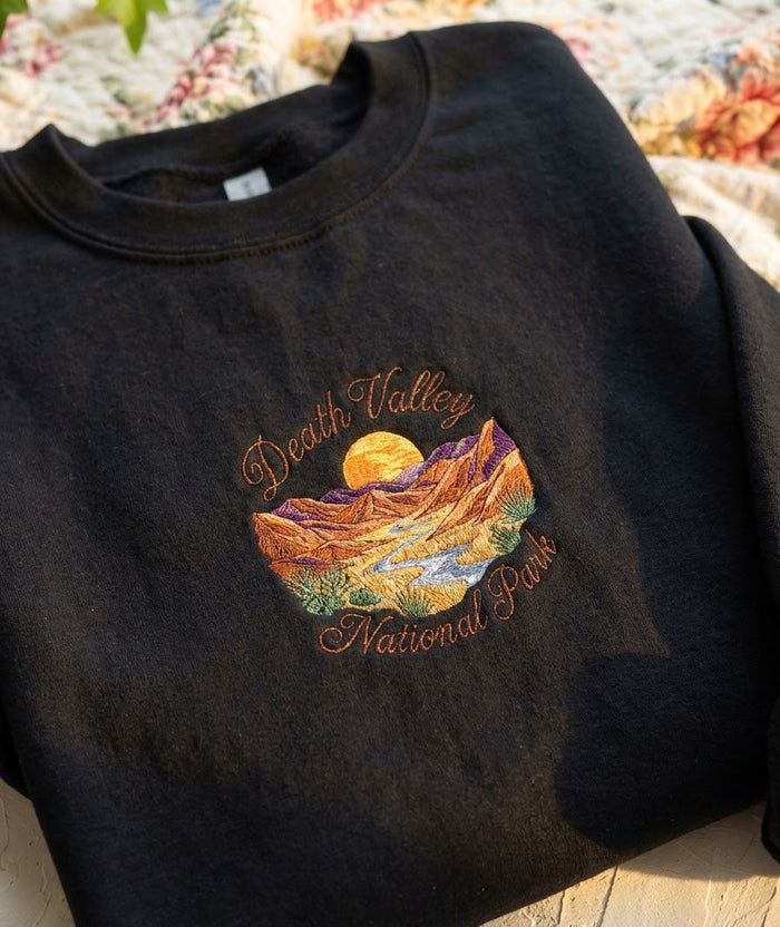 A folded black crewneck sweatshirt featuring a colorful embroidered 'Death Valley National Park' graphic on the chest, displayed on a light wooden surface with a floral blanket in the background.