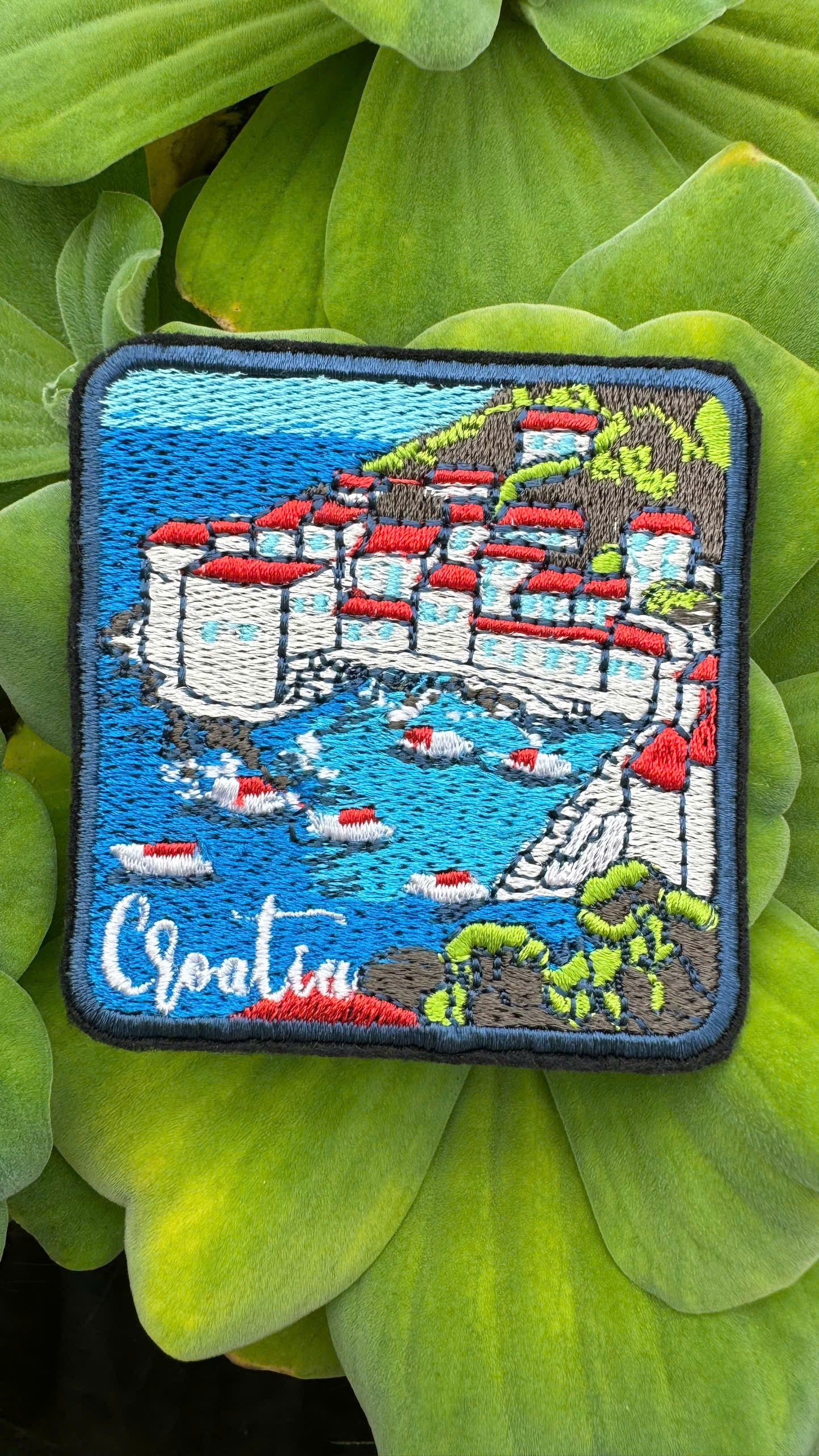 Embroidered patch of a coastal scene with buildings and boats on a green leaf background