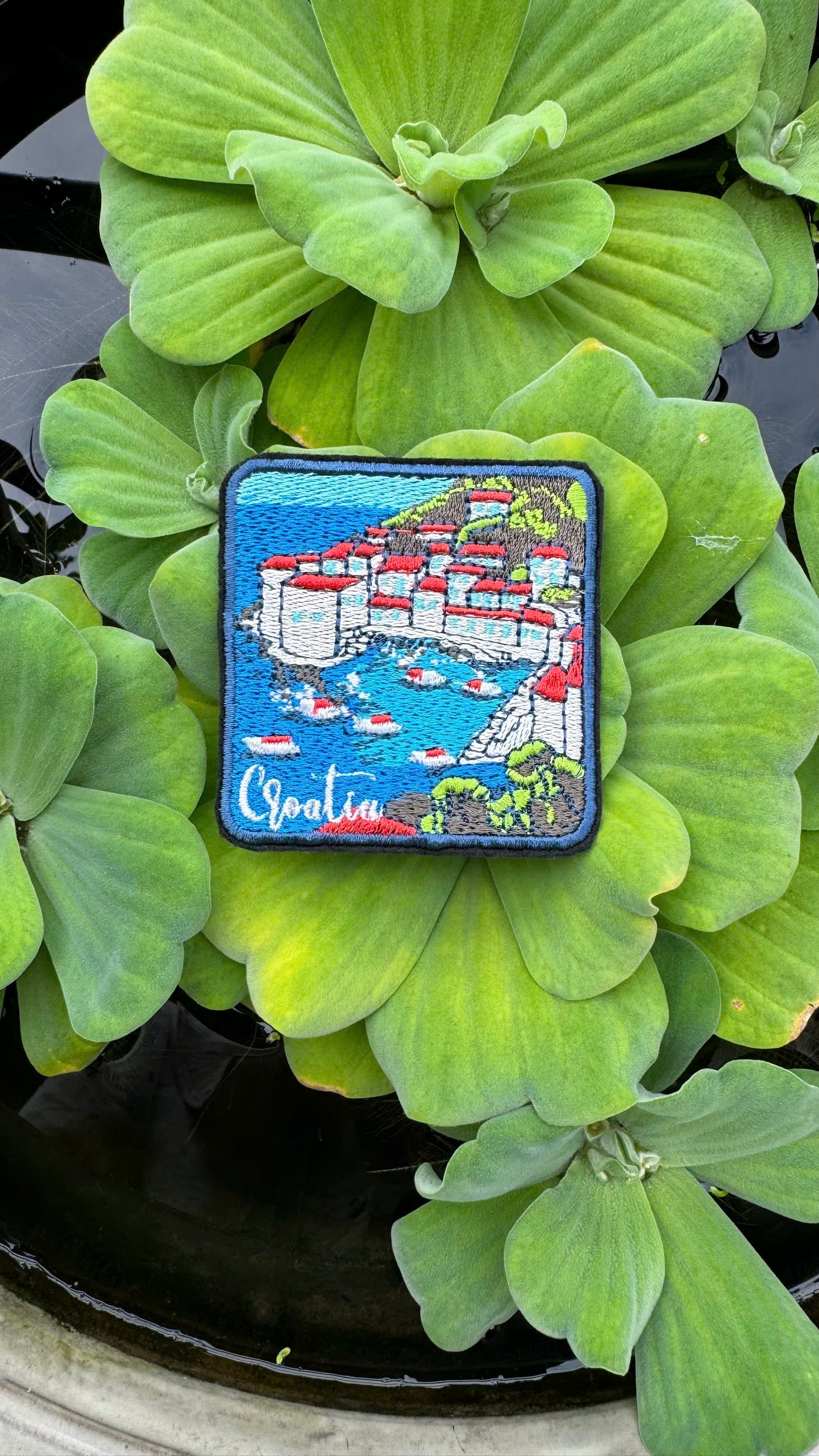 Embroidered patch of a coastal scene with buildings and boats, placed on green leaves.