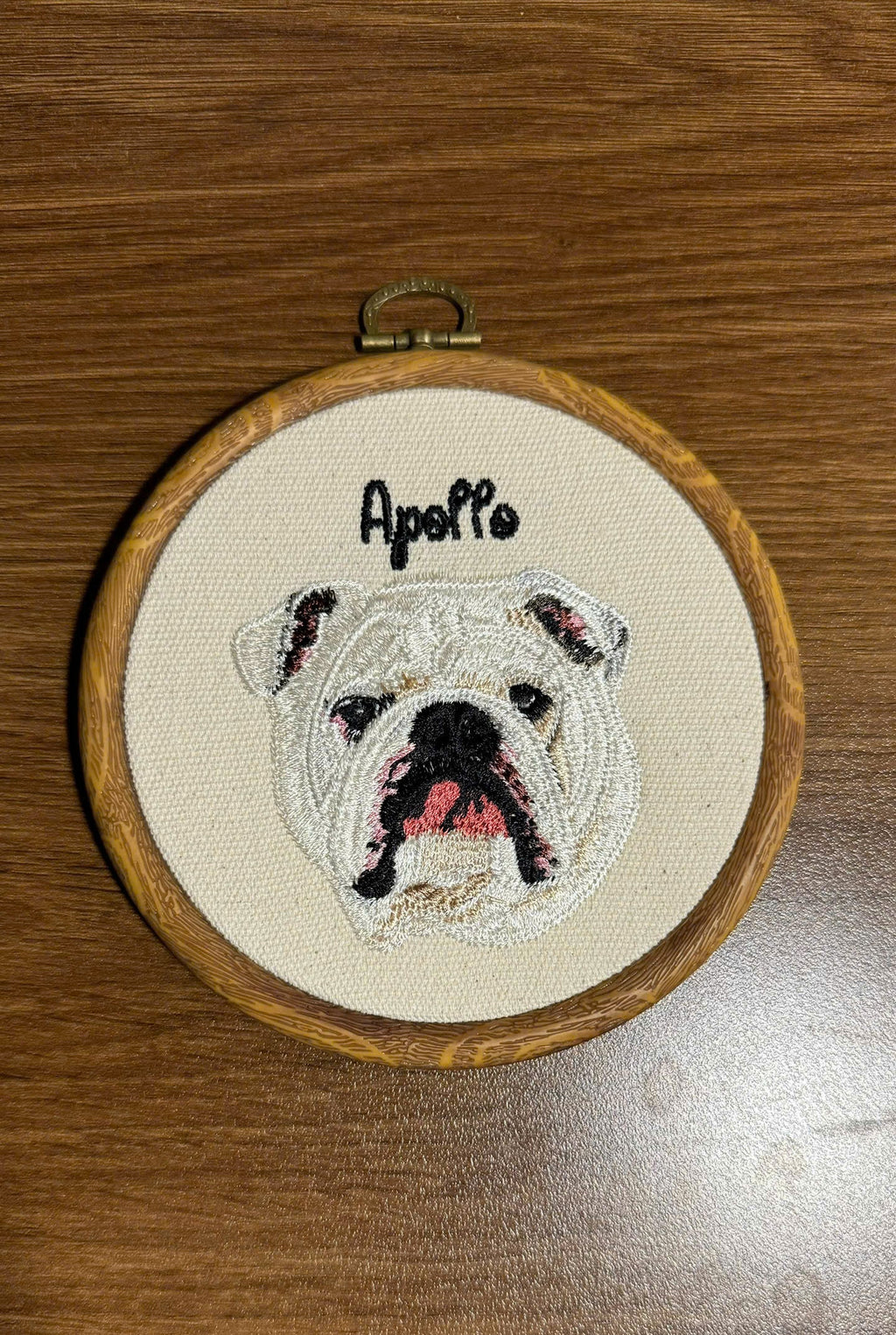 Embroidery of a dog's face with 'Apollo' above it in a wooden hoop on a wooden surface.