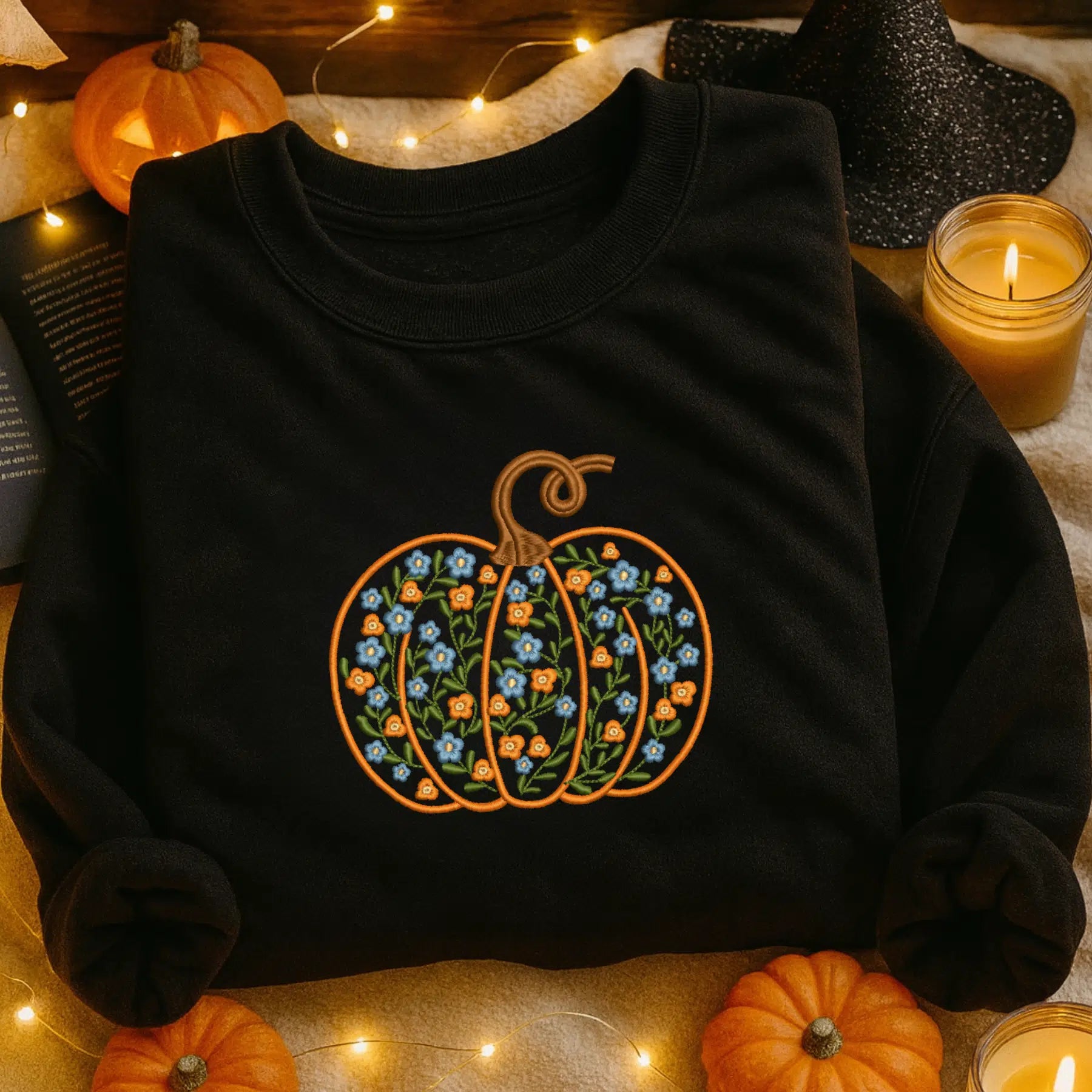 Black sweatshirt with floral pumpkin design on a Halloween-themed background with pumpkins and candles.