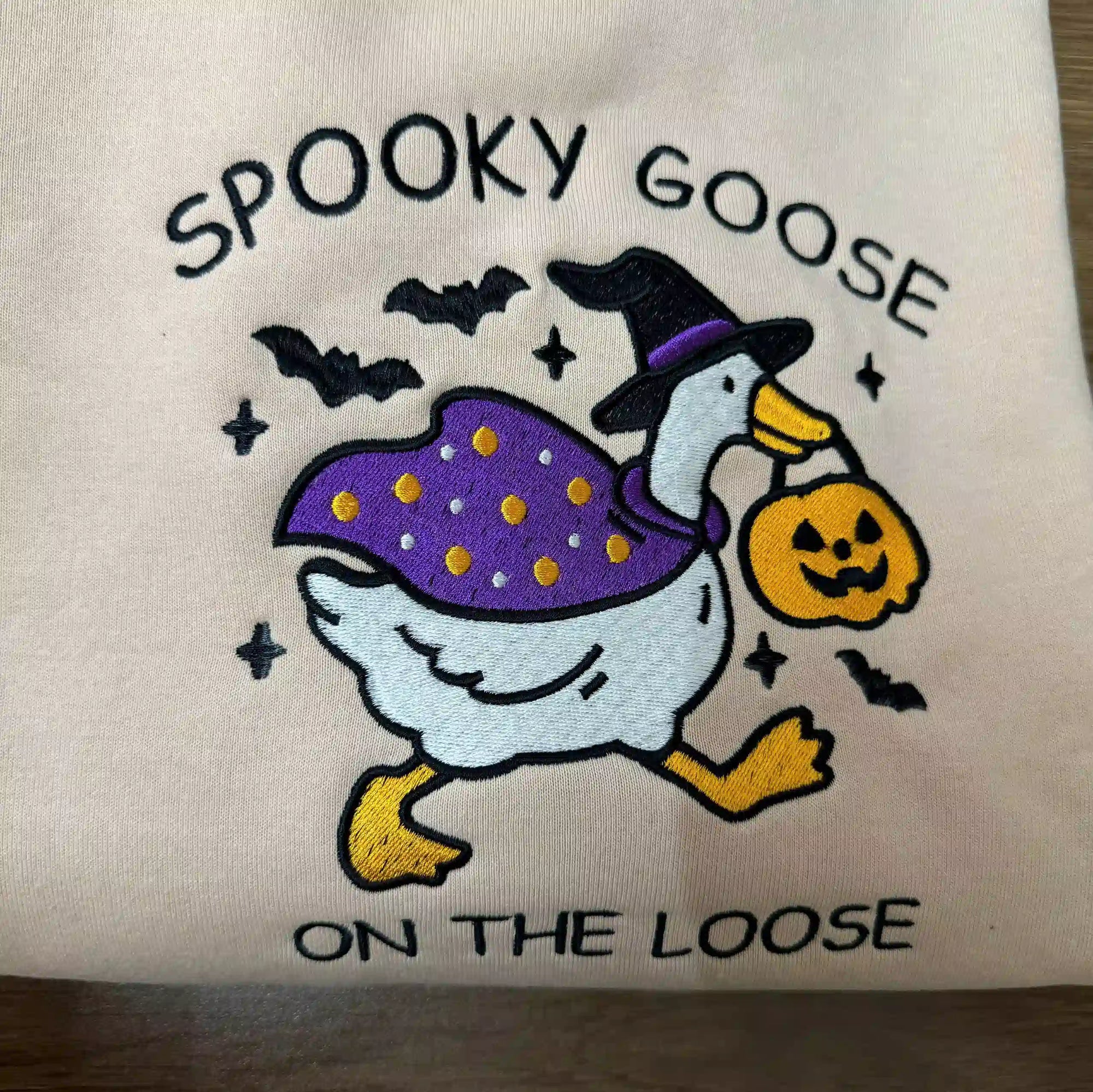 Embroidered design of a goose in a witch costume with 'Spooky Goose on the Loose' text on a beige background.