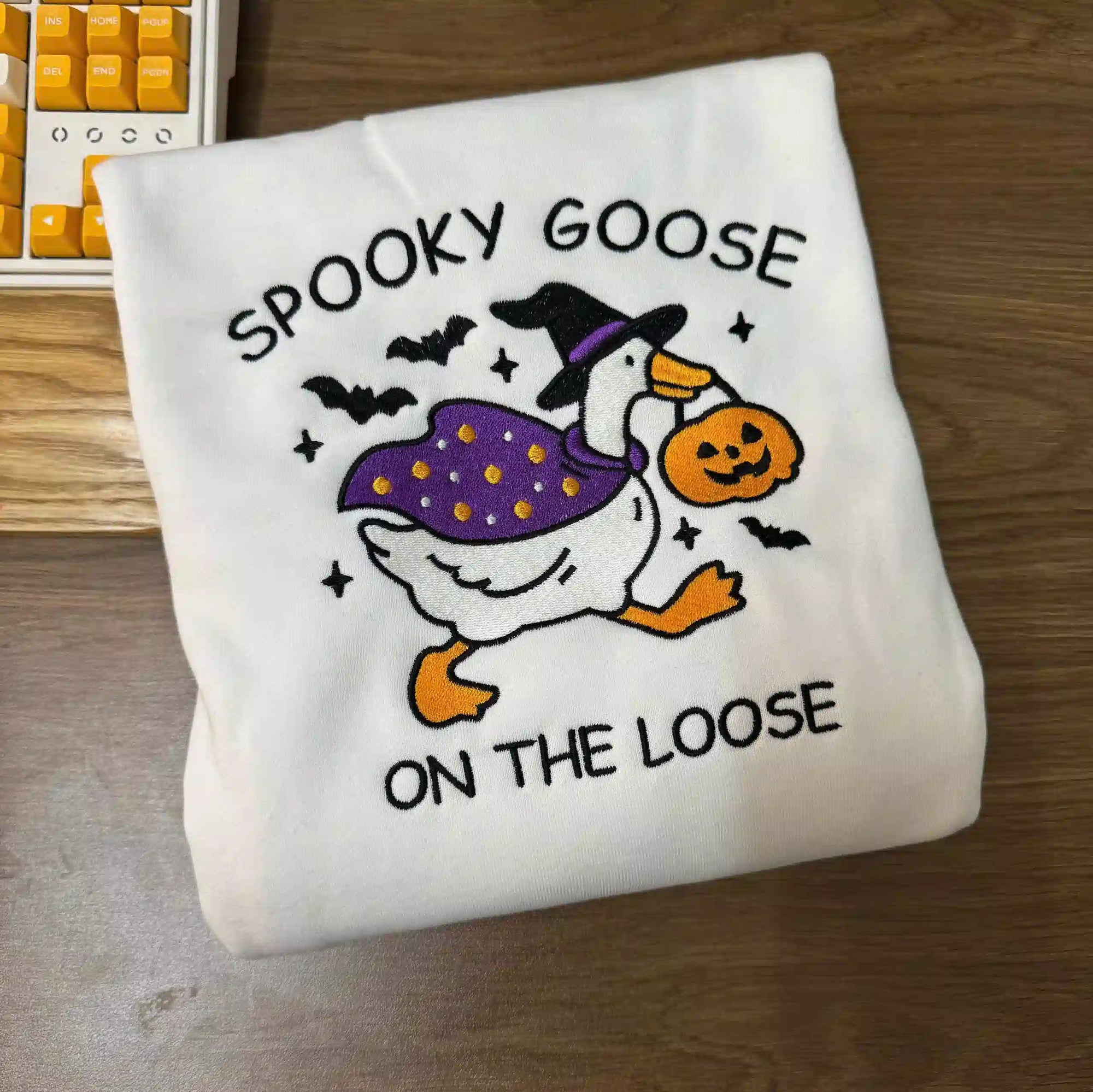 White sweatshirt with Halloween-themed design of a goose and text on a wooden surface.