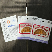 Two packages of 'Le Croissant' patches with a thank you card on a wooden surface.