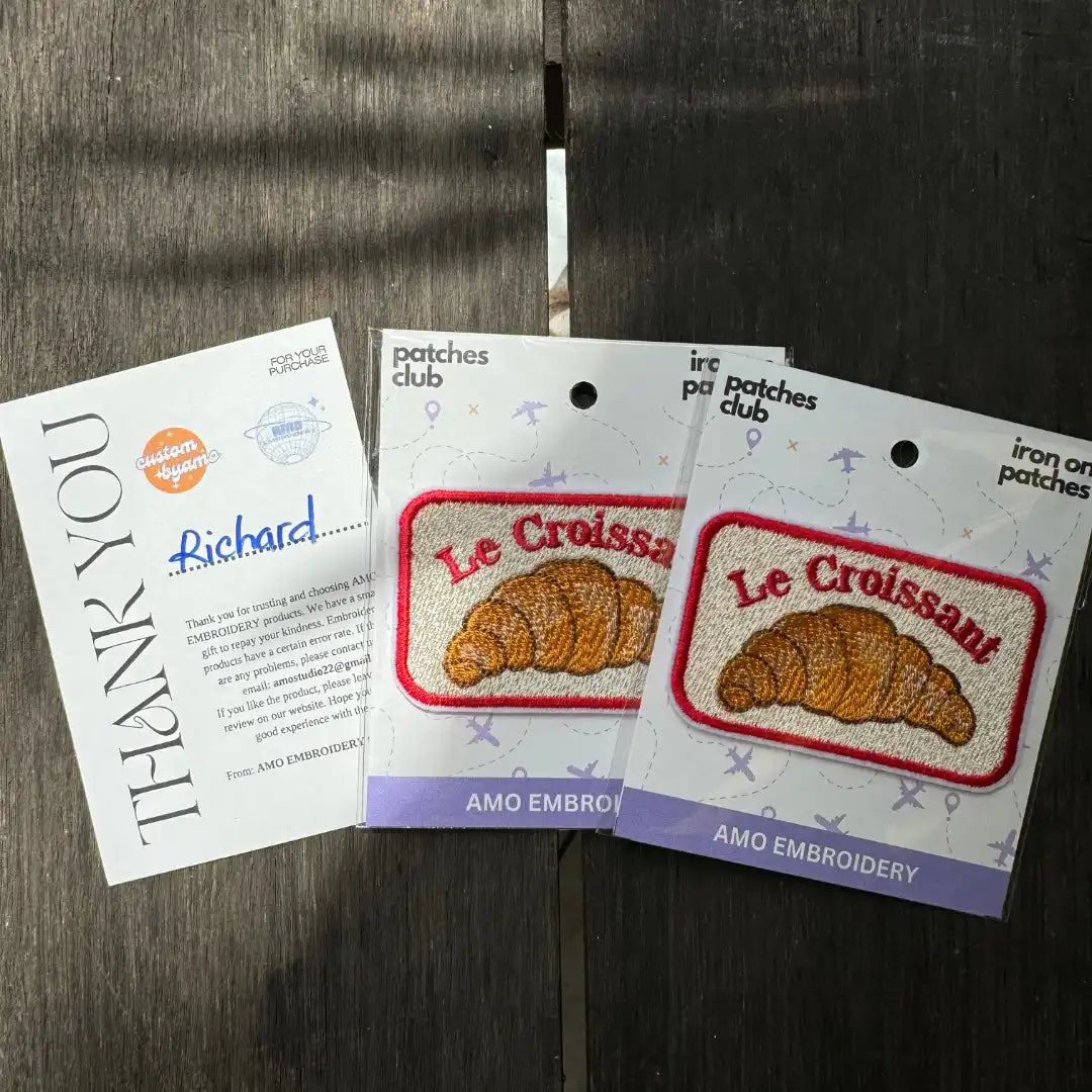 Two packages of 'Le Croissant' patches with a thank you card on a wooden surface.