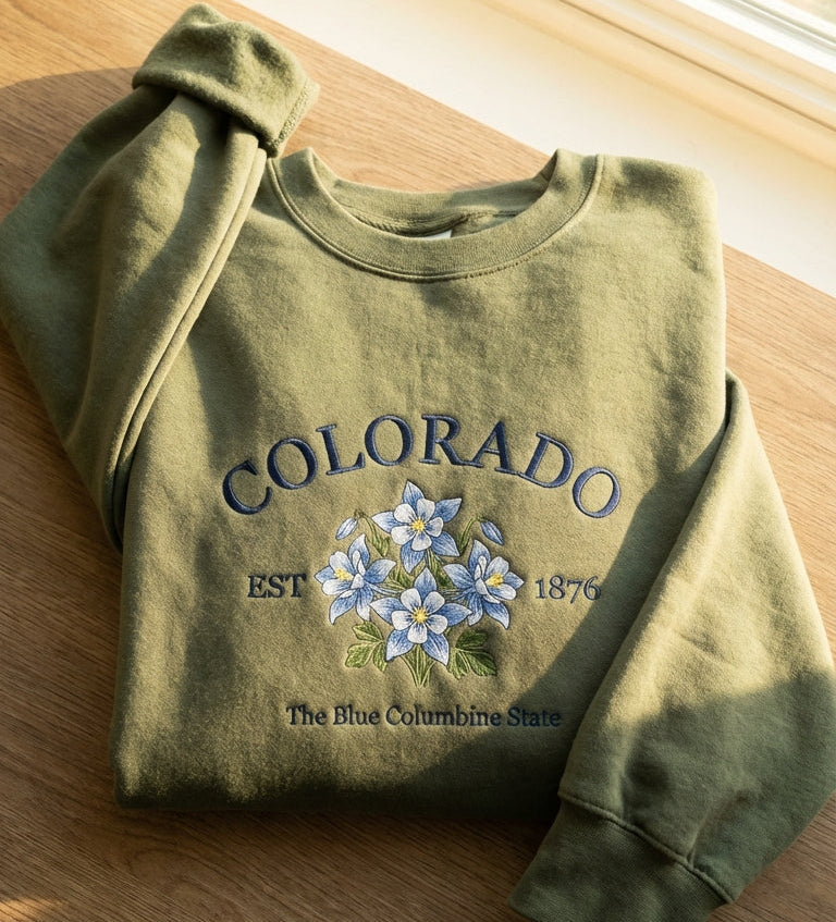 Olive green folded sweatshirt with detailed blue and white Columbine flower embroidery reading Colorado EST 1876 The Blue Columbine State