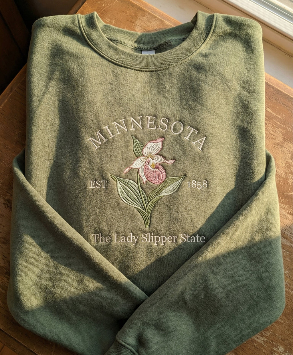 Olive green folded sweatshirt with detailed pink and white Lady Slipper orchid embroidery reading Minnesota EST 1858 The Lady Slipper State