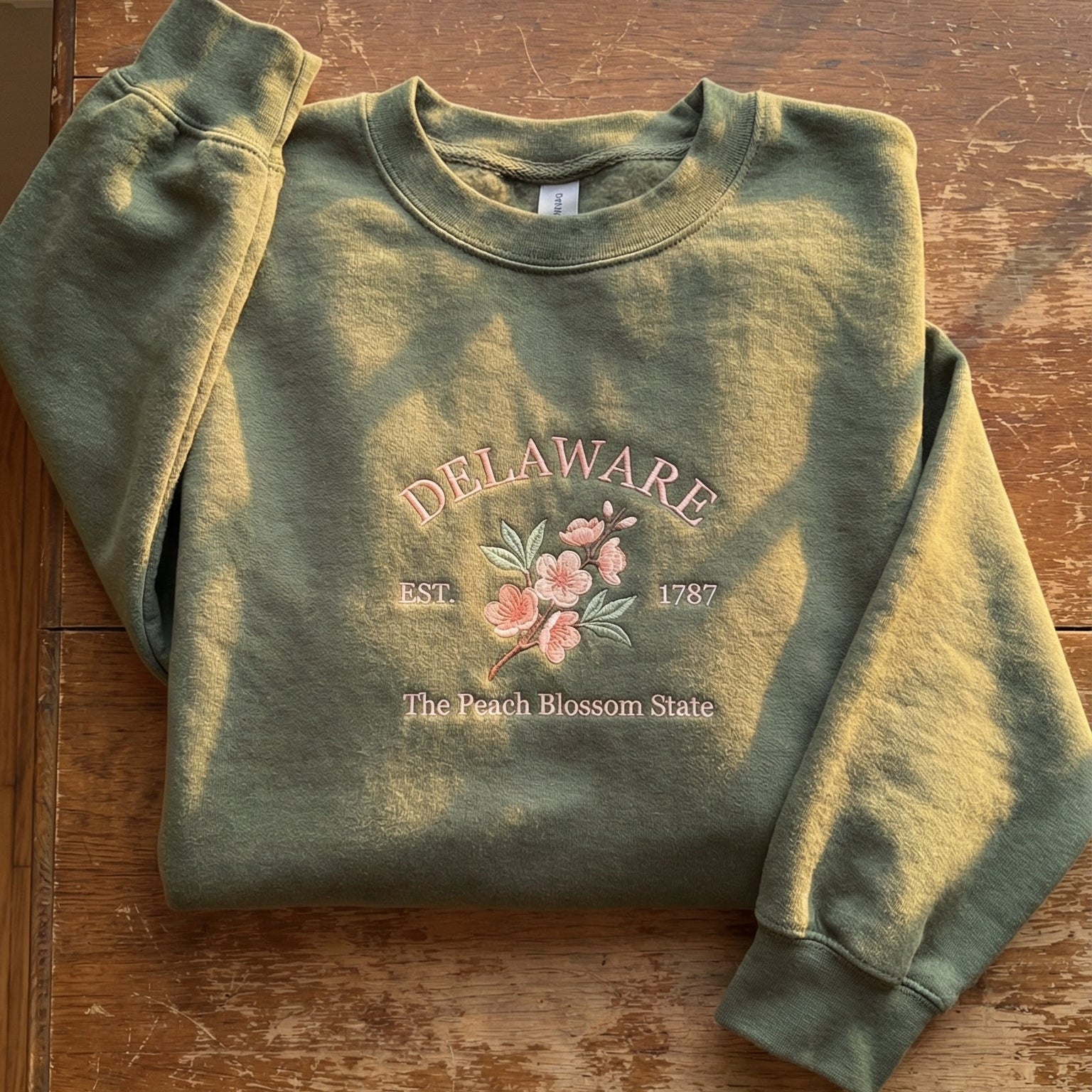 Olive green sweatshirt with detailed pink Peach Blossom flower embroidery reading Delaware EST. 1787 The Peach Blossom State