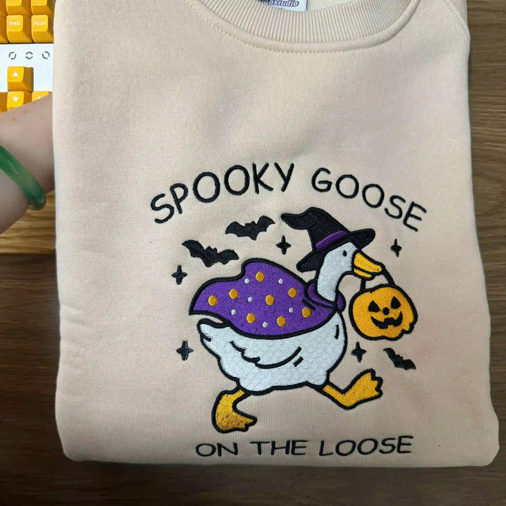 Folded beige sweatshirt with 'Spooky Goose on the Loose' design featuring a goose in a witch hat and pumpkin bag.