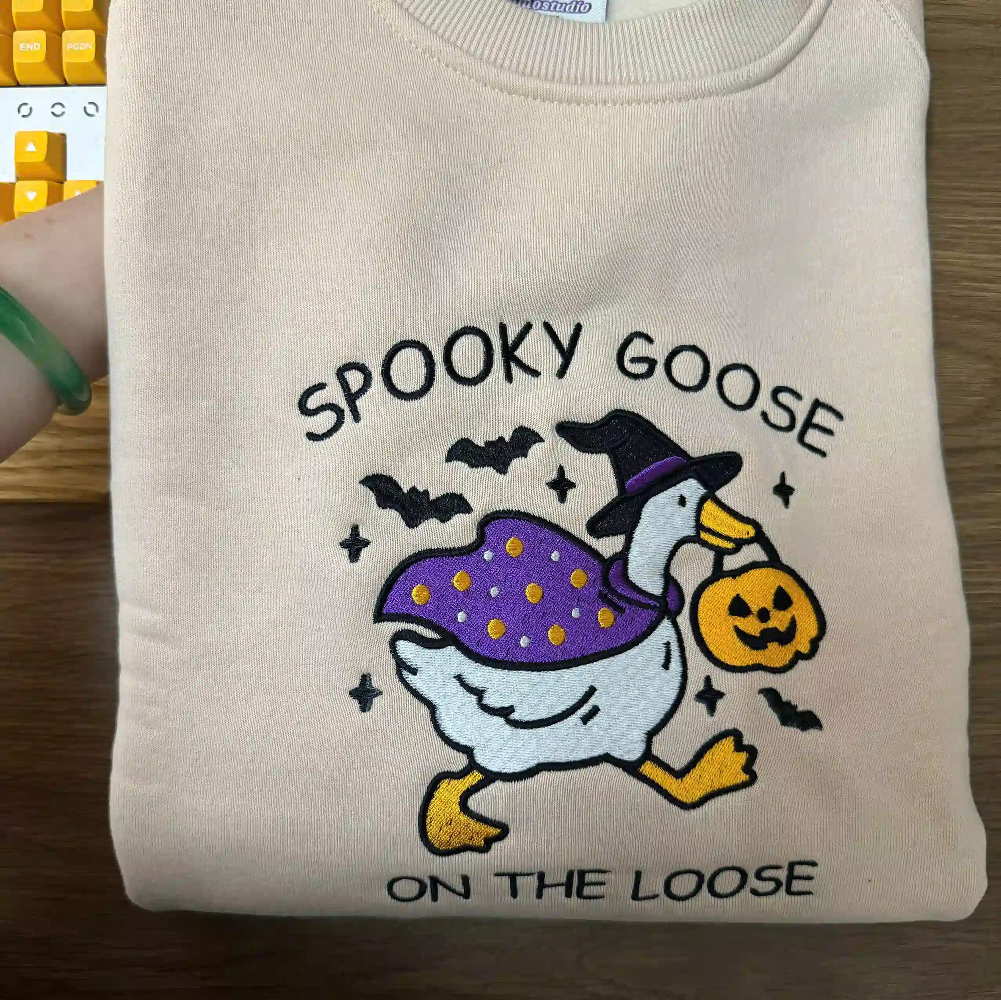 Folded beige sweatshirt with 'Spooky Goose on the Loose' design featuring a goose in a witch hat and pumpkin bag.