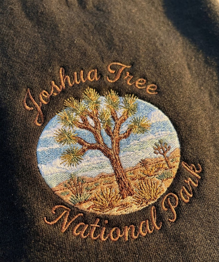 Close-up of a dark brown embroidered patch featuring a Joshua Tree National Park design with a central circular desert scene and the words 'Joshua Tree National Park' in brown script.