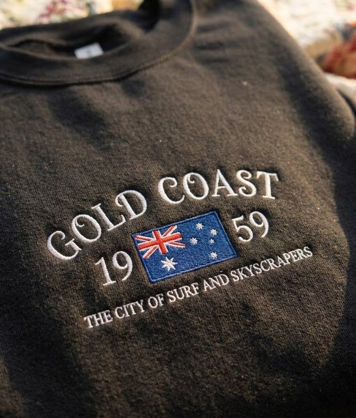A close-up of a dark charcoal gray t-shirt featuring an embroidered graphic on the chest. The design reads 'GOLD COAST 1959' in white, with a small Australian flag in the center and 'THE CITY OF SURF AND SKYSCRAPERS' embroidered below.