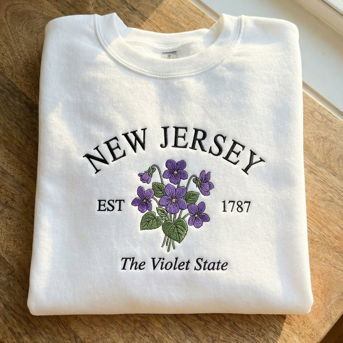 White folded sweatshirt with detailed Purple Lilac flower embroidery reading New Hampshire EST 1788 The Purple Lilac State