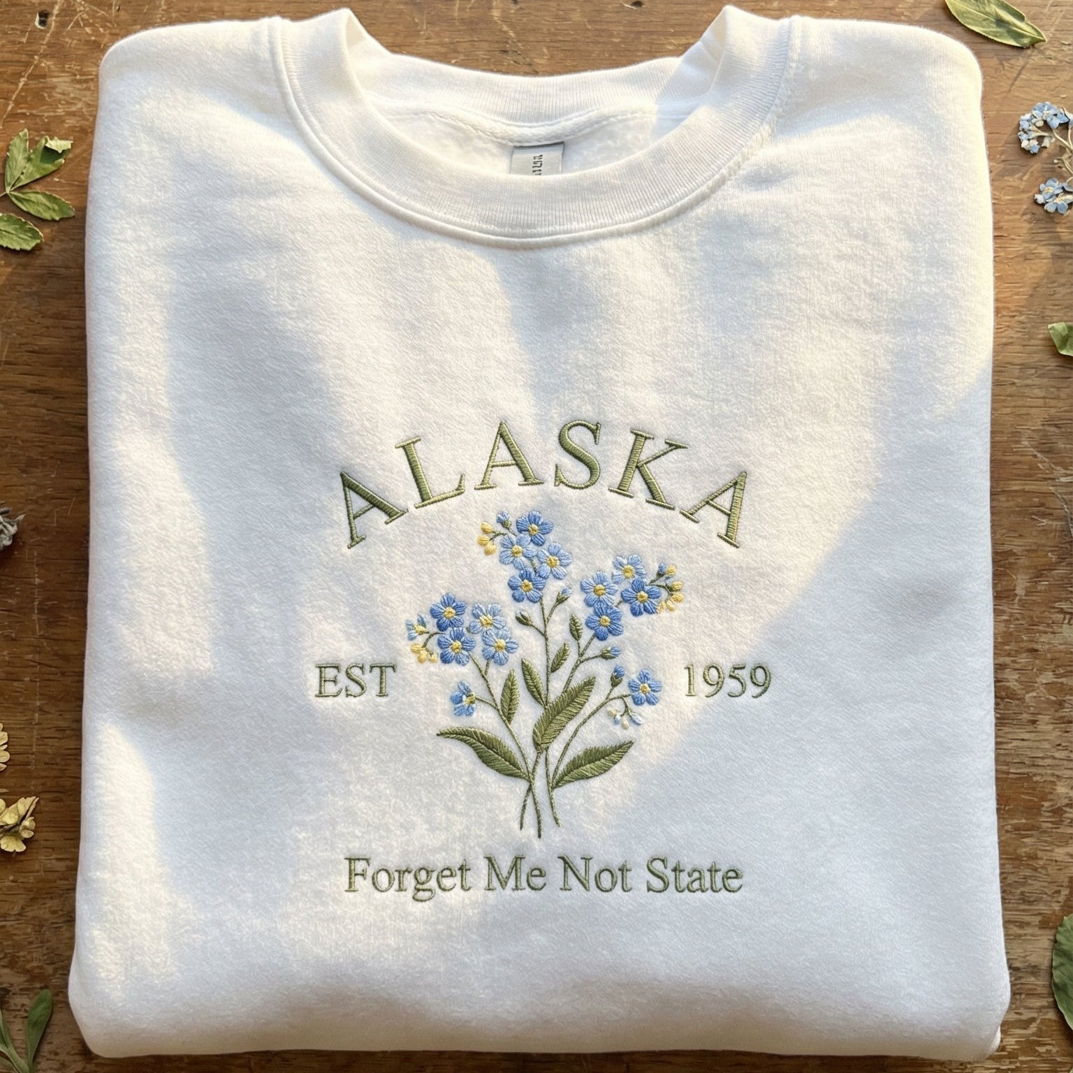 White folded sweatshirt with detailed blue Forget-Me-Not flower embroidery reading Alaska EST 1959 Forget Me Not State