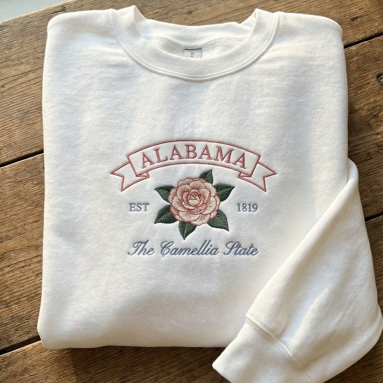 White folded sweatshirt with detailed pink Camellia flower embroidery and a pink banner reading Alabama EST 1819 The Camellia State