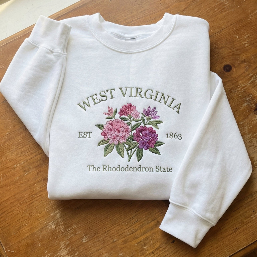 White folded sweatshirt with detailed pink and purple Rhododendron flower embroidery, reading West Virginia EST 1863 The Rhododendron State