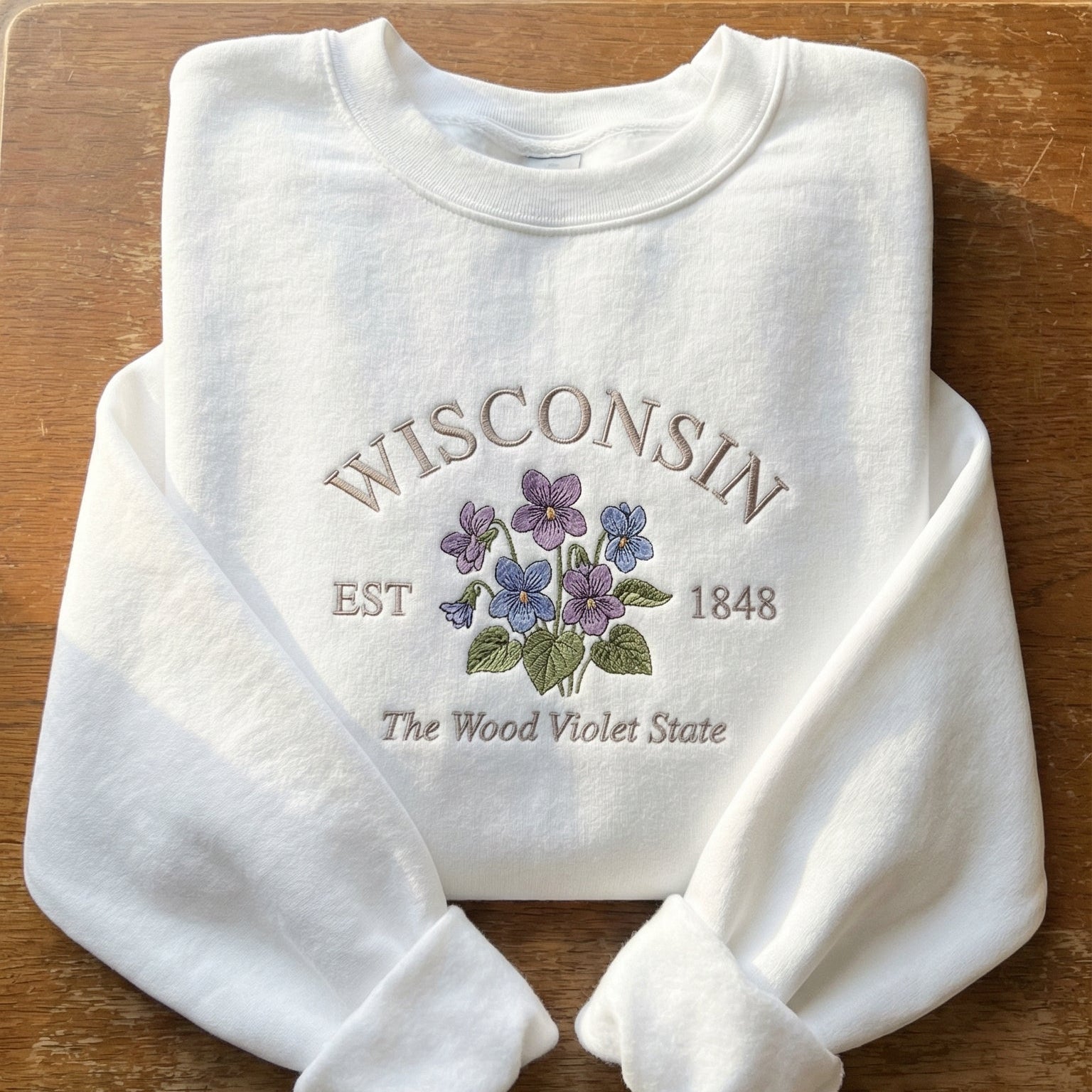 White folded sweatshirt with detailed purple Wood Violet flower embroidery reading Wisconsin EST 1848 The Wood Violet State