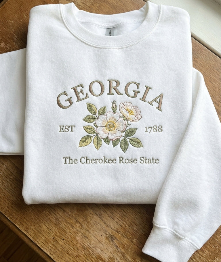 White folded sweatshirt with detailed white and gold Cherokee Rose flower embroidery reading Georgia EST 1788 The Cherokee Rose State