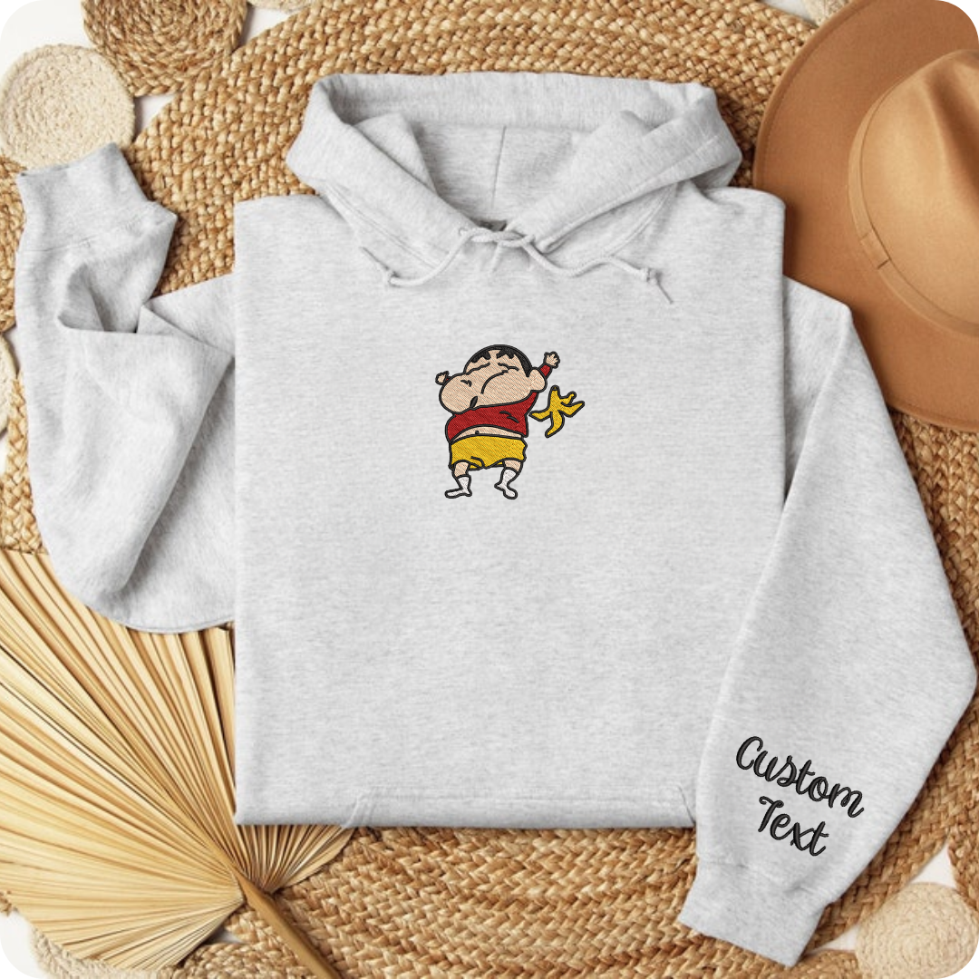 Light gray embroidered hoodie with jumping ShinChan character, flat lay on woven texture